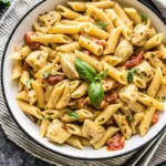 A bowl of creamy penne pasta with chunks of chicken, sun-dried tomatoes, and garnished with fresh basil leaves, served on a striped napkin with a fork and knife on the side